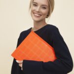 A cheerful woman smiles while holding an orange folder against a neutral background.
